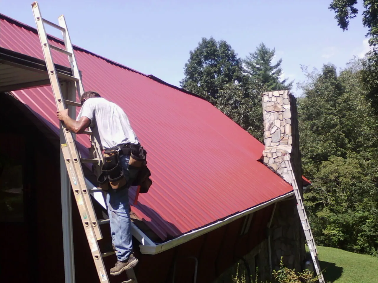 Roofer on a ladder beside a red metal roof and stone chimney — Soffit & Fascia Repair by Roofing Brothers in Blanch